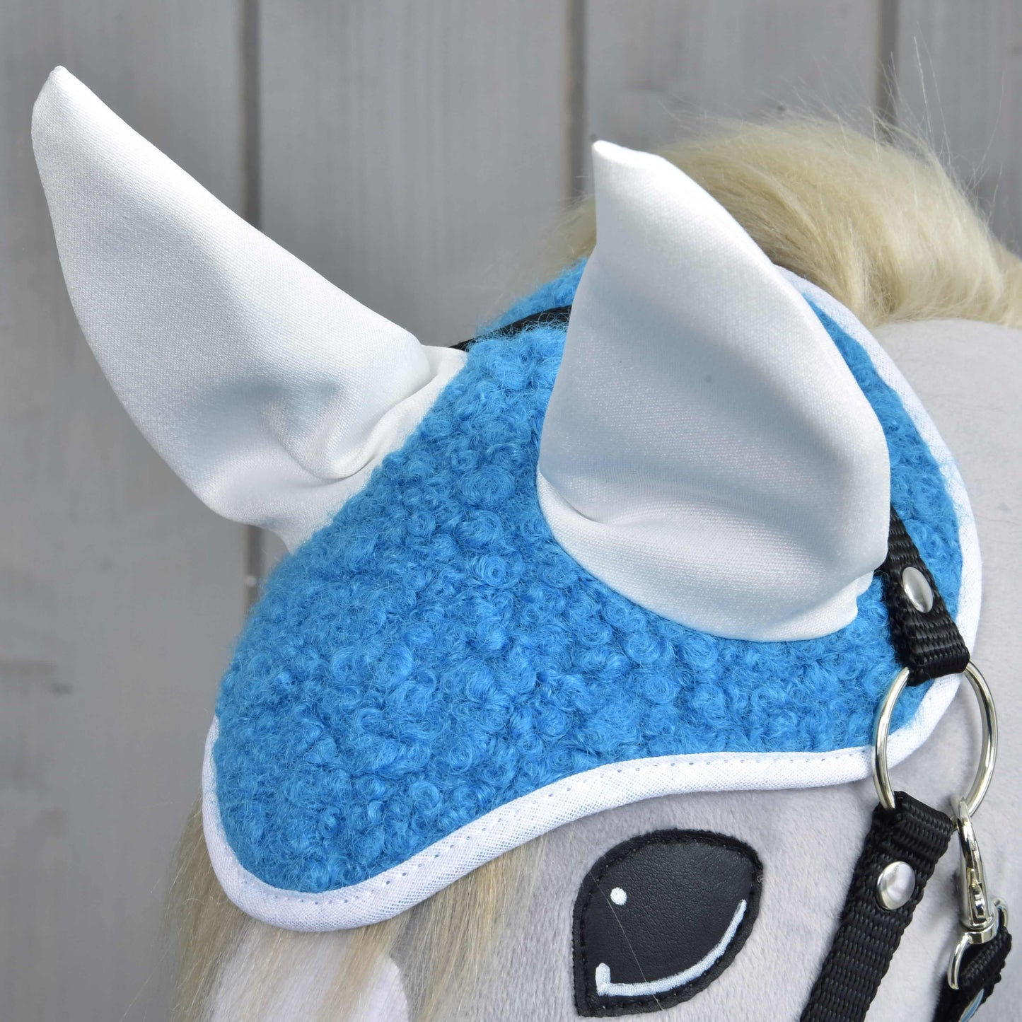 Nahaufnahme von Hobby Horse LarDen Set Mystery mit einem Spielzeugpferdekopf mit blau-weißer Fliegenhaube, großen aufrechten Ohren, Zaumzeug und comicartigen Augen vor hellem Holzhintergrund – ideal als charmante Dekoration.