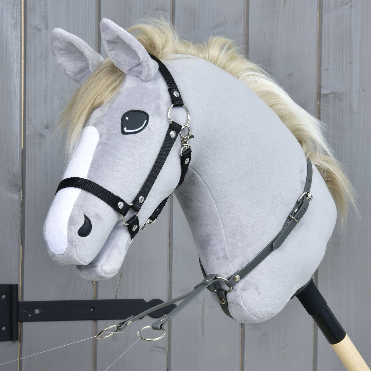 Nahaufnahme eines plüschigen Hobby Horse LarDen „Martingale dunkelgrau“ mit weißer Mähne, schwarzem Zaumzeug und Kunstleder-Martingal, auf einem Holzstab vor einer grauen Holzwand.