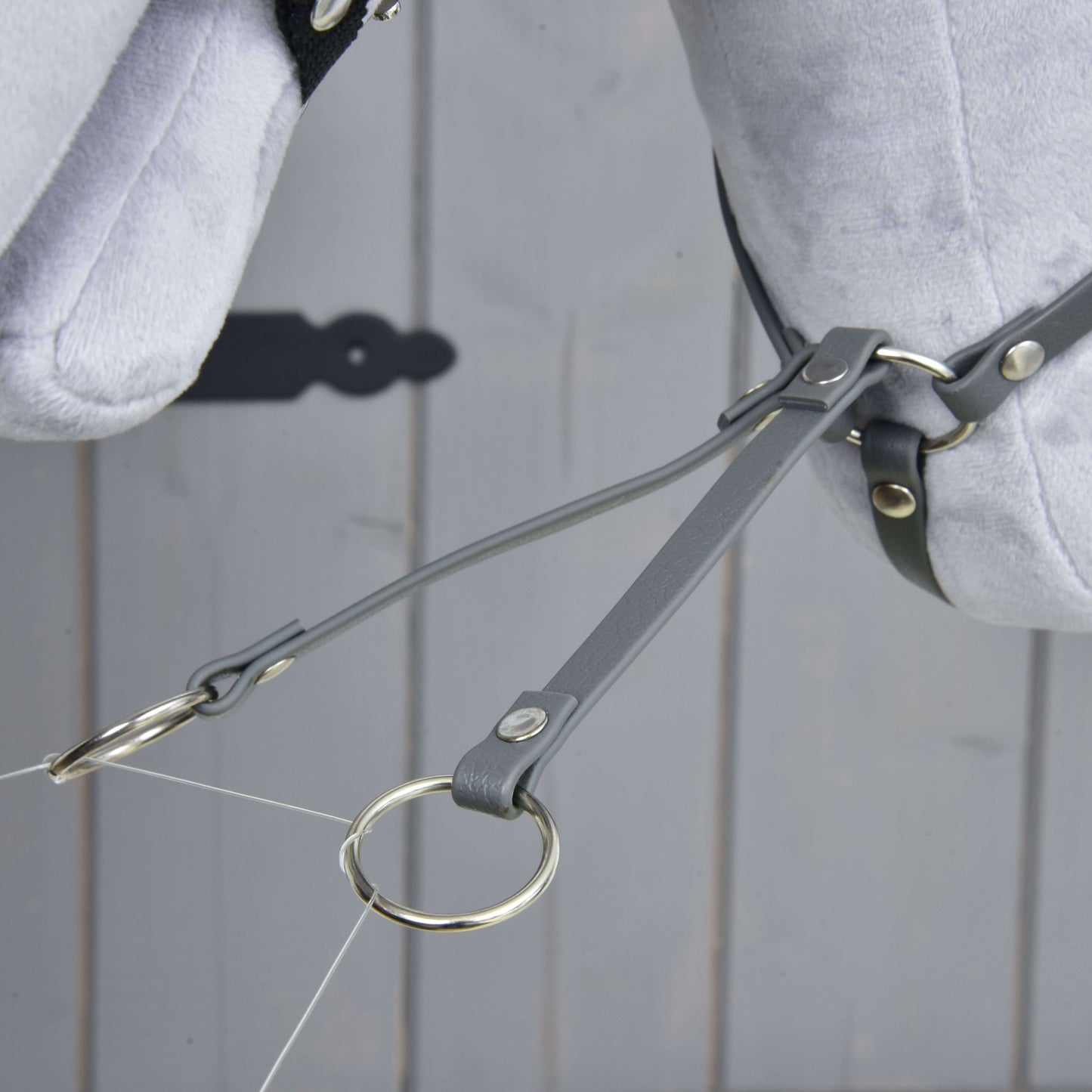 Nahaufnahme der Martingale dunkelgrau von Hobby Horse LarDen mit grauen Lederriemen und einem Kunstleder-Martingal mit Metallringen an einem hellgrauen Plüschpferdekopf vor Holz-Hintergrund.