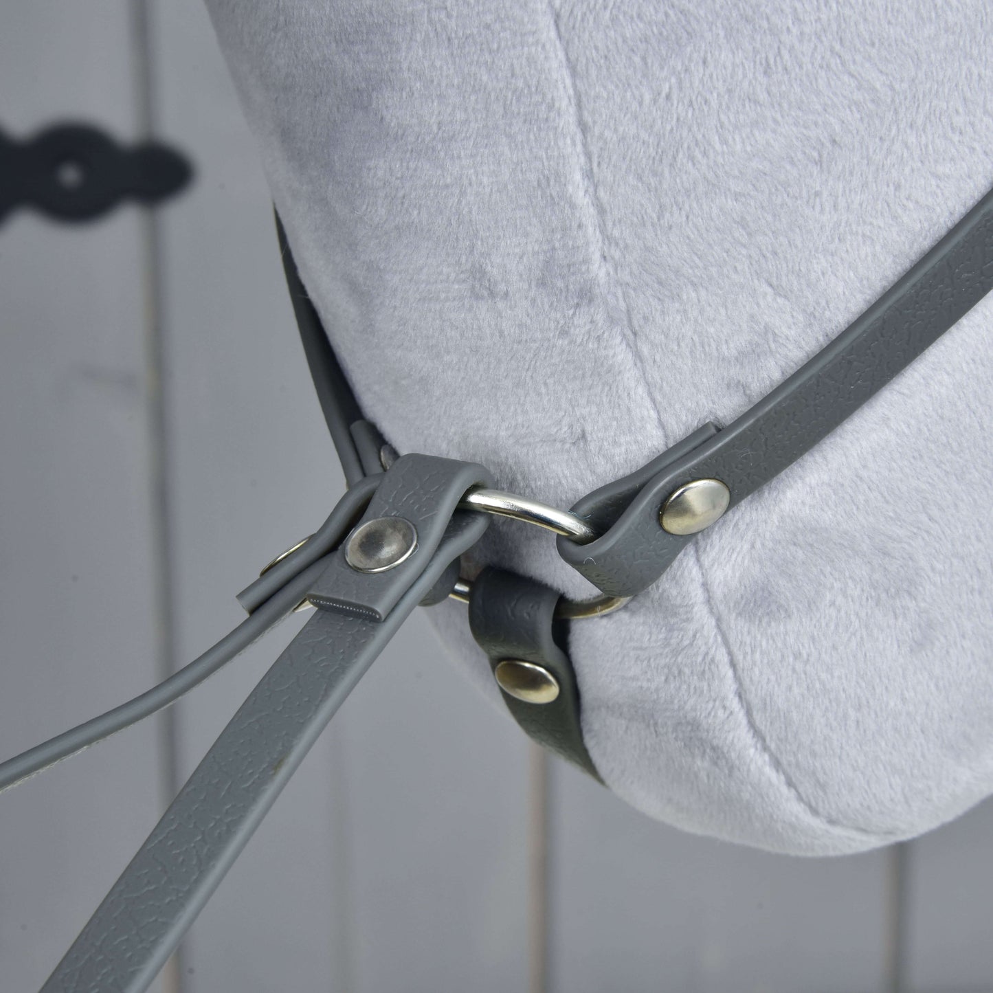 Nahaufnahme der Martingale dunkelgrau von Hobby Horse LarDen an einem hellgrauen Plüschpferdekopf mit grauen Lederriemen und Metallringen vor verschwommenem Holz-Hintergrund.