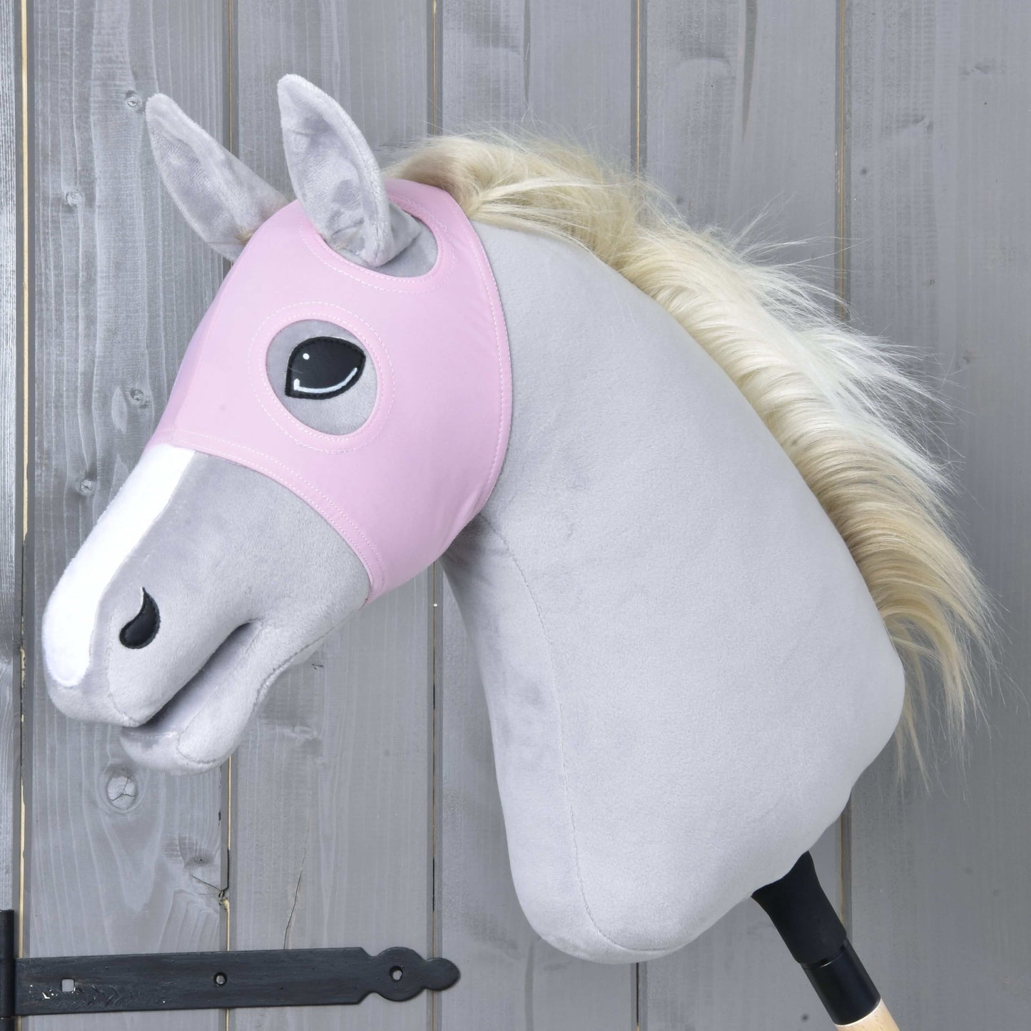 Ein Plüsch-Hobby Horse von Hobby Horse LarDen trägt Maske ohne Ohren Pink, eine waschbare rosa Maske, auf seinem hellgrauen Körper mit weißer Schnauze und cremefarbener Mähne. Das Pferd ist auf einem Holzstab montiert vor einer grauen Holzwand.