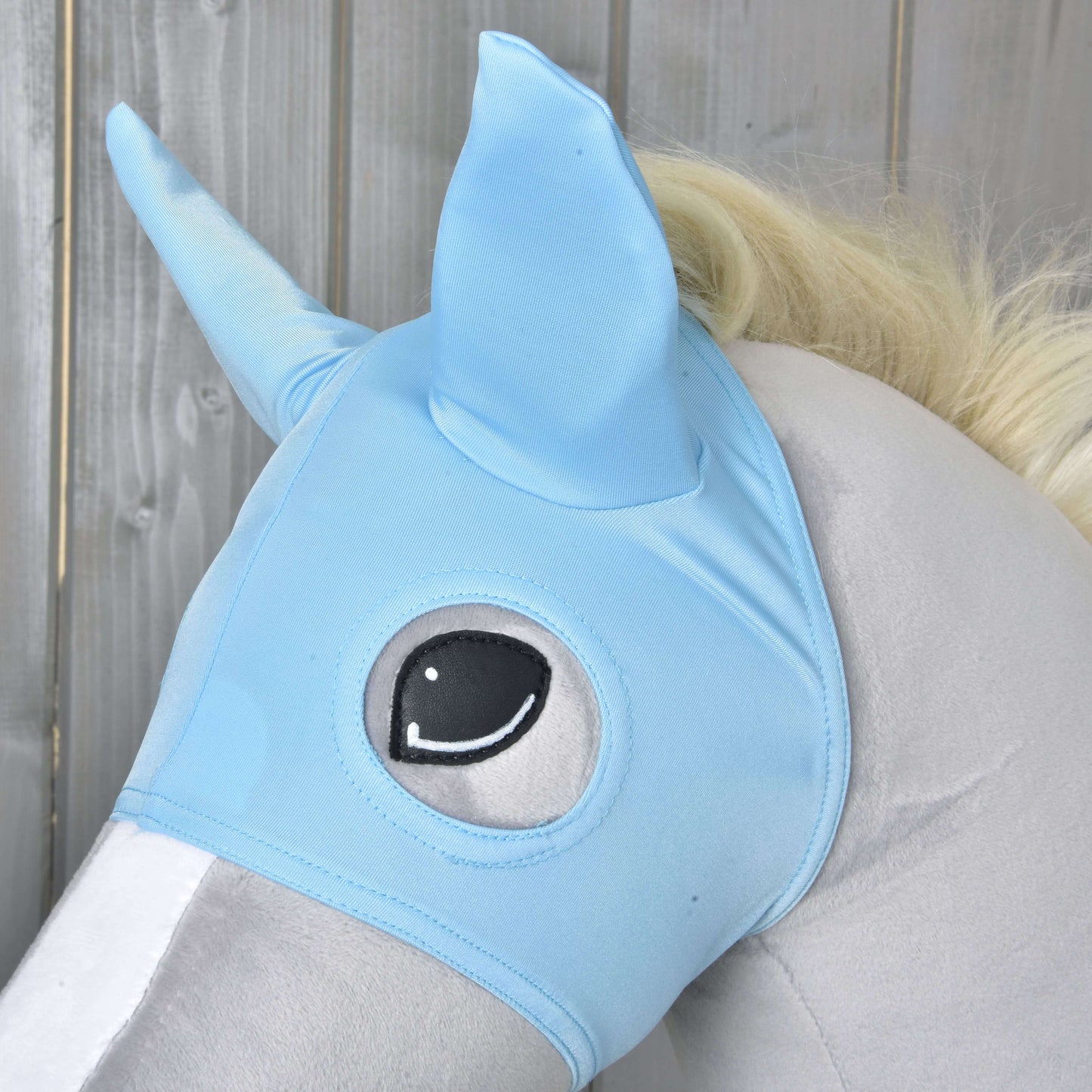 Gros plan sur un cheval-jouet en peluche portant le Masque de Cheval Ciel de Hobby Horse LarDen, avec des oreilles pointues et un œil illustré, sur fond en bois.