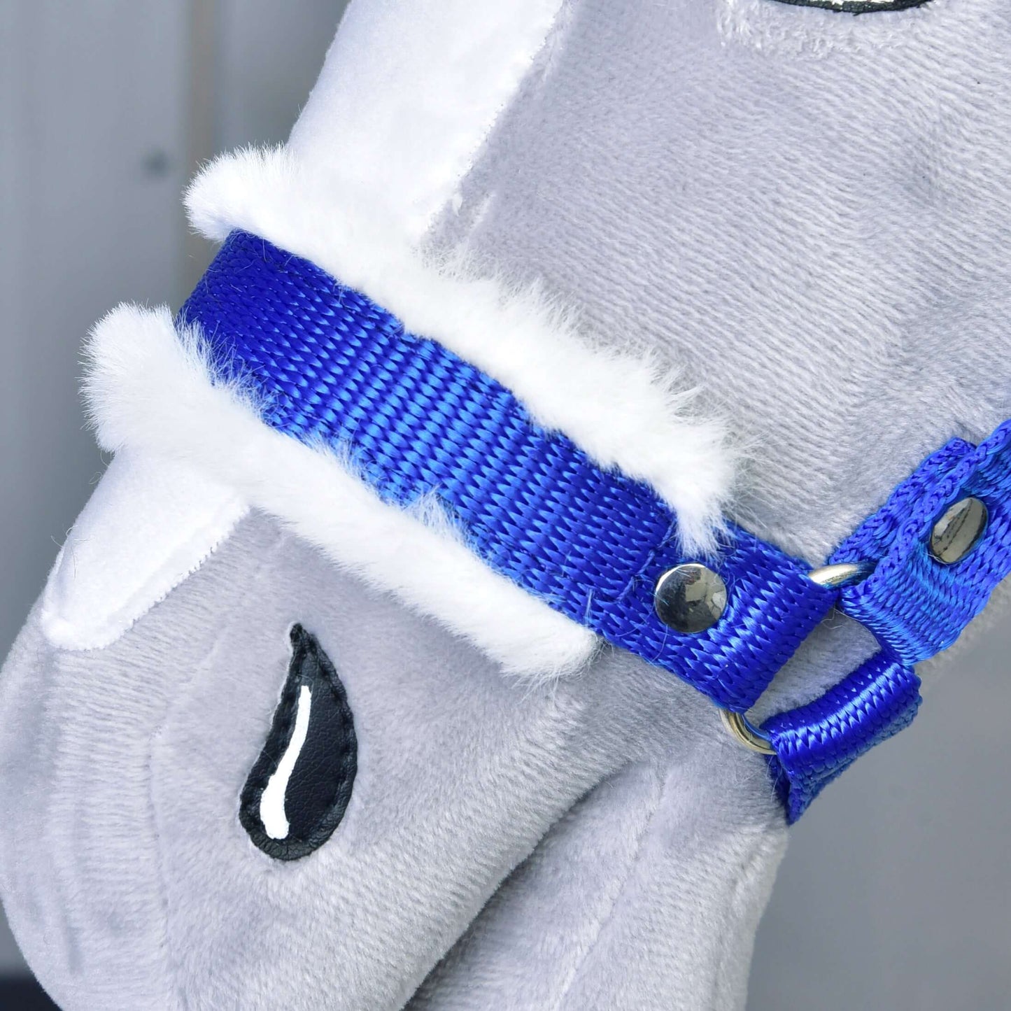 Gros plan sur le visage d'un cheval en peluche Hobby Horse LarDen, mettant en valeur le Licol bleu avec fourrure. Détails : bordure en fausse fourrure blanche, yeux noirs brodés avec détail en goutte sur tissu gris clair.