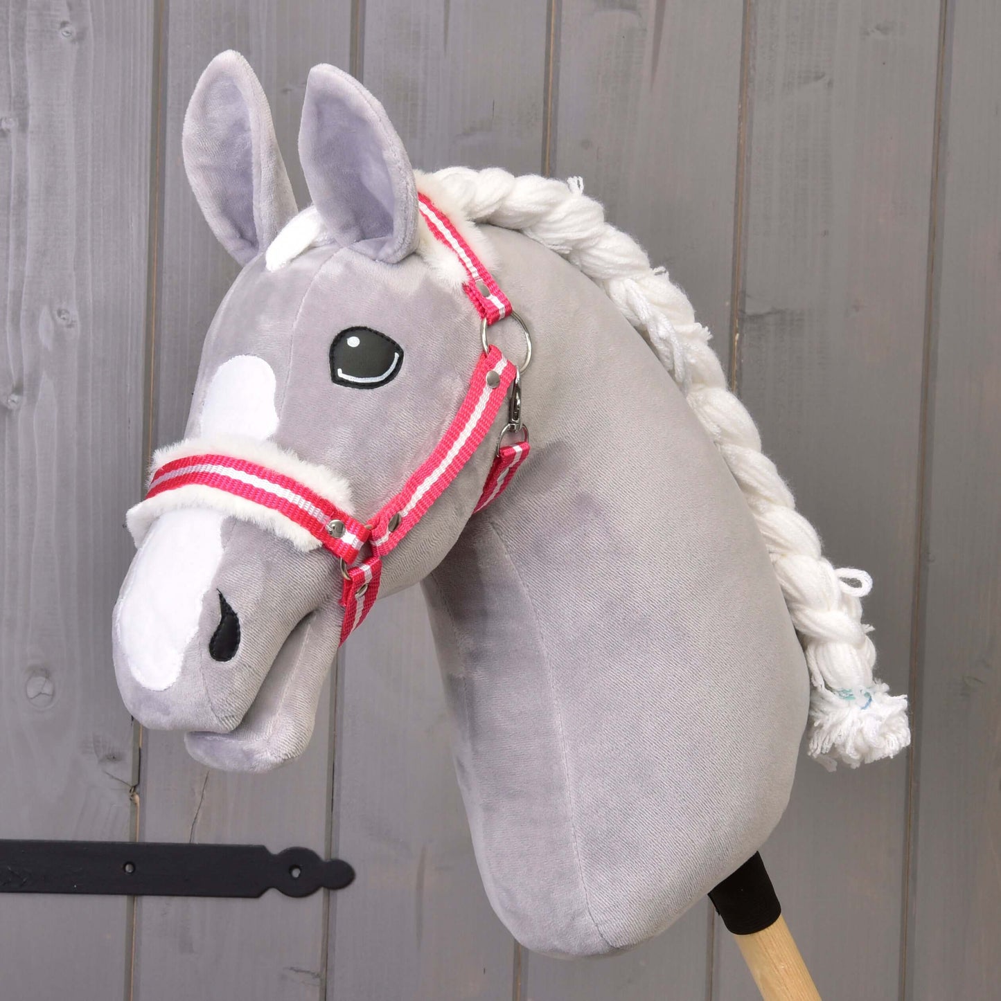 Licol rayé rouge avec fourrure de Hobby Horse LarDen sur un Hobby Horse gris en peluche avec crinière blanche et licol à rayures rouges, monté sur un bâton en bois devant un mur en bois gris.