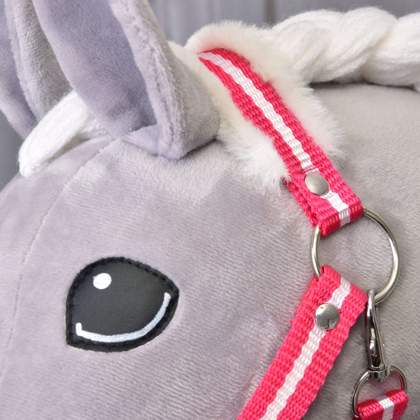 Gros plan sur une tête de licorne grise en peluche avec crinière blanche, œil brodé noir et blanc, et le Licol rayé rouge avec fourrure de Hobby Horse LarDen.