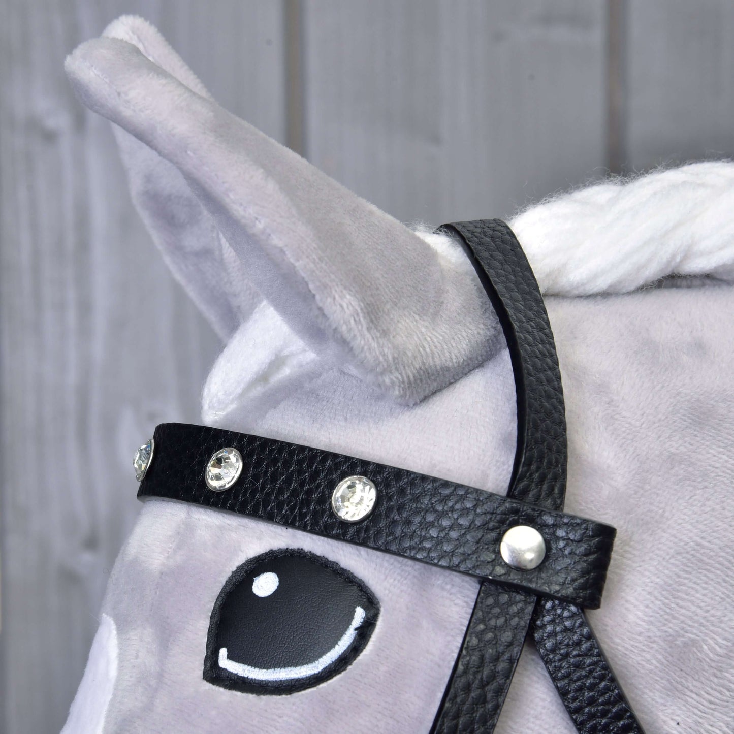 Nahaufnahme eines grauen Plüschpferdekopfes mit Komplettes Kunstlederzaumzeug mit Ziersteinen von Hobby Horse LarDen. Das Pferd hat eine weiße Mähne, ein aufgerichtetes Ohr und ein comicartiges Auge, im Hintergrund helle Holzplanken.