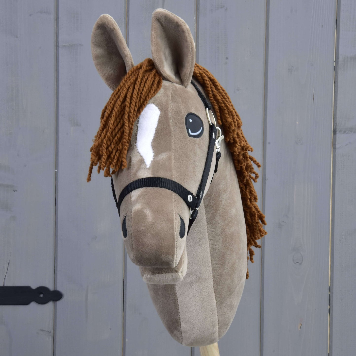 Hobby Horse Oliver avec licol noir + licol en fourrure GRATUIT de Hobby Horse LarDen, une peluche d’étalon Oldenbourg à fourrure brune, avec liste blanche, crinière en fil brun et licol noir, incluant un licol en fourrure offert. Présenté devant un fond en bois gris.