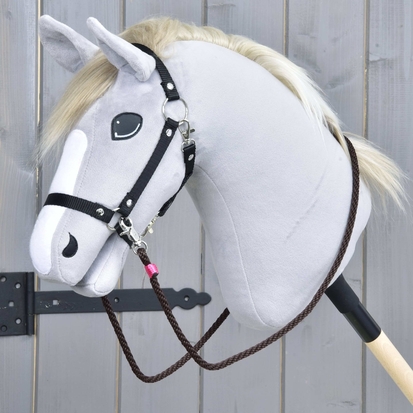 Gros plan sur l’ensemble de corde Noir Cacao de Hobby Horse LarDen, un Hobby Horse gris en peluche avec bride noire, licol et rênes marron sur un bâton en bois, sur fond en bois clair.
