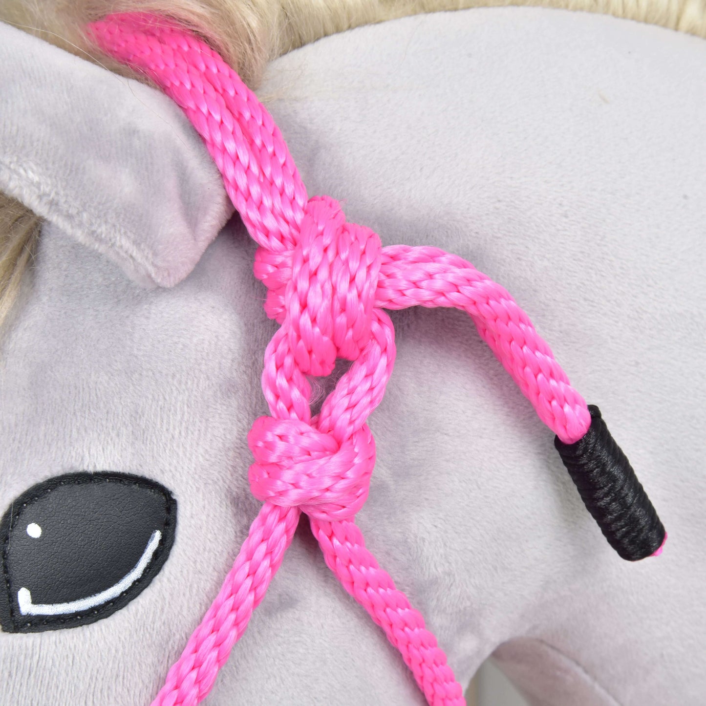 Close-up of a plush horse's head wearing the Hobby Horse LarDen Rope halter Pink, tied around its ear. The horse features a light gray body, painted black eye with white highlights, and a light blonde mane.