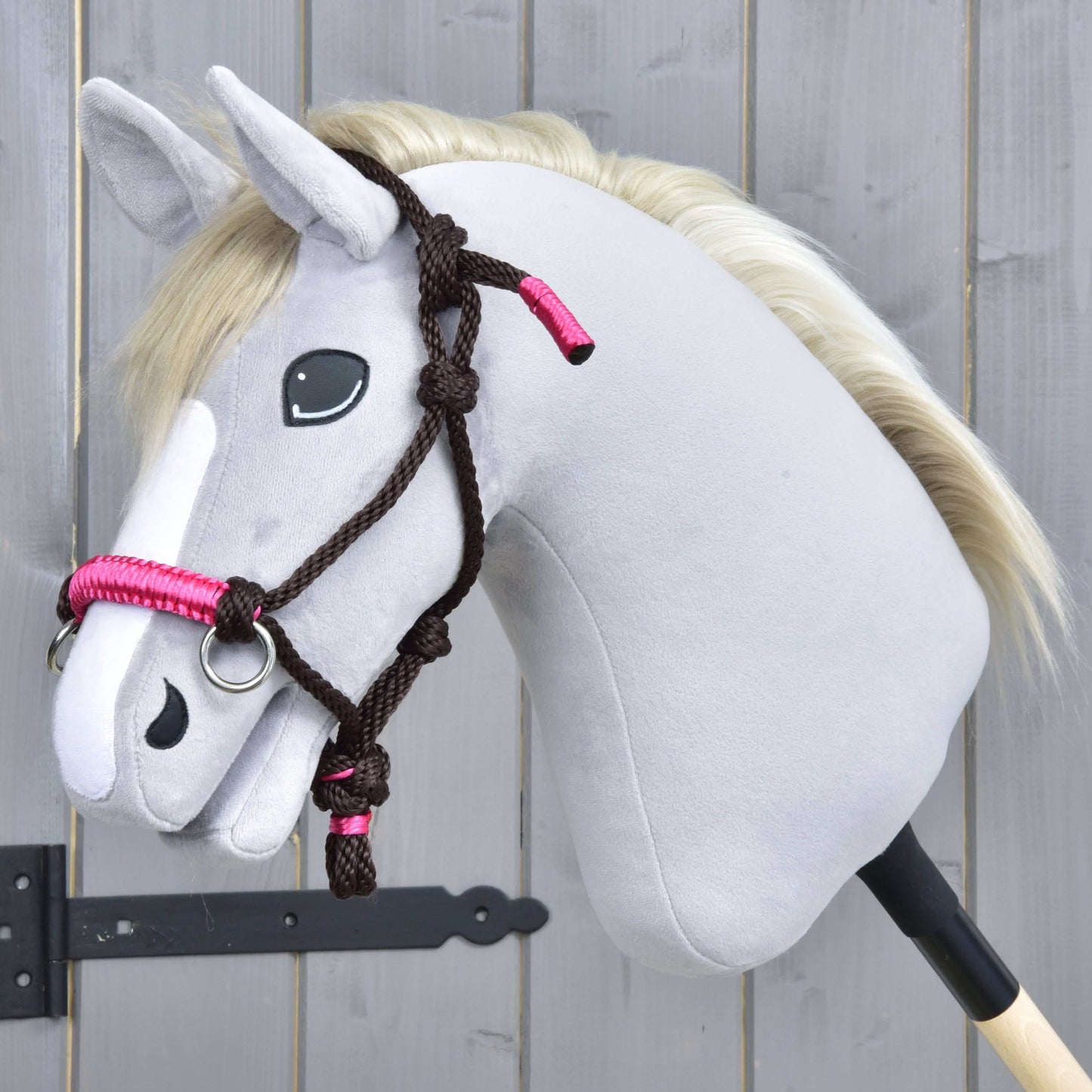 Ensemble de corde Noir Cacao de Hobby Horse LarDen, Hobby Horse gris en peluche sur bâton avec bride noire et rose, rênes, yeux noirs en bouton et crinière blanche, présenté devant une clôture en bois.