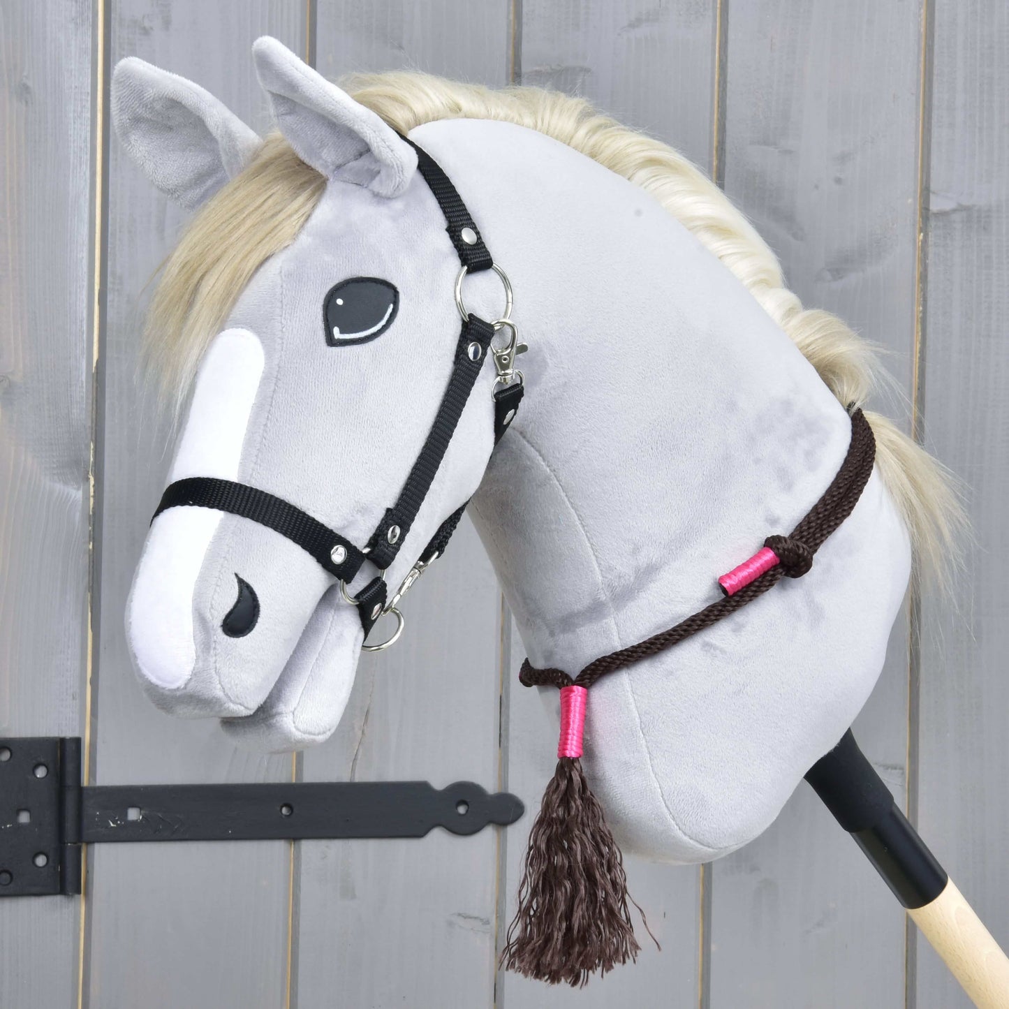 Ensemble de corde Noir Cacao de Hobby Horse LarDen, Hobby Horse gris en peluche avec crinière blanche, bride noire et rênes en corde marron avec accents roses sur un bâton en bois, posé contre un fond en bois gris.