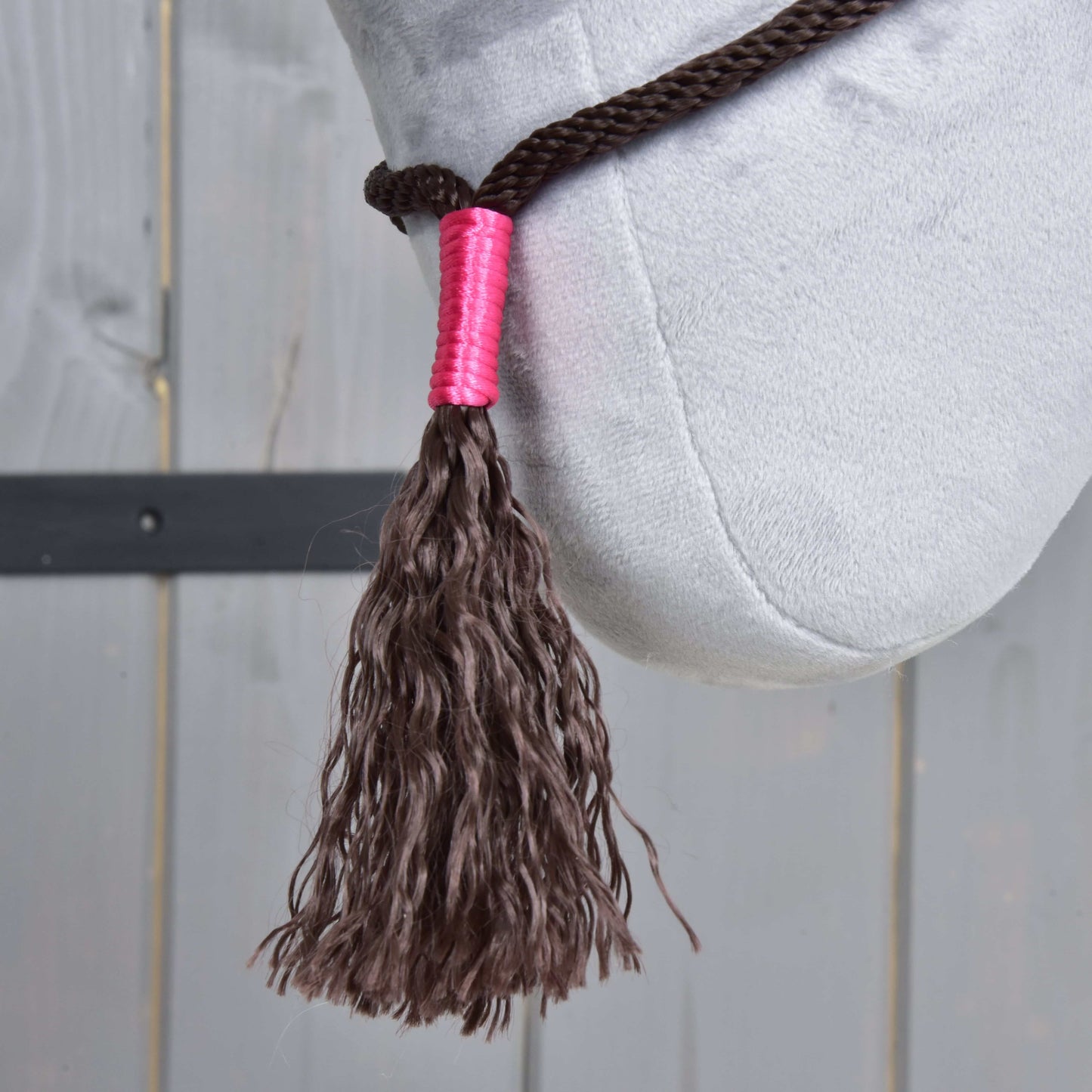 Gros plan sur l’ensemble de corde Noir Cacao de Hobby Horse LarDen montrant un pompon tressé marron foncé sur des rênes épaisses avec enroulement rose, présenté sur une surface en peluche gris clair avec fond en bois.