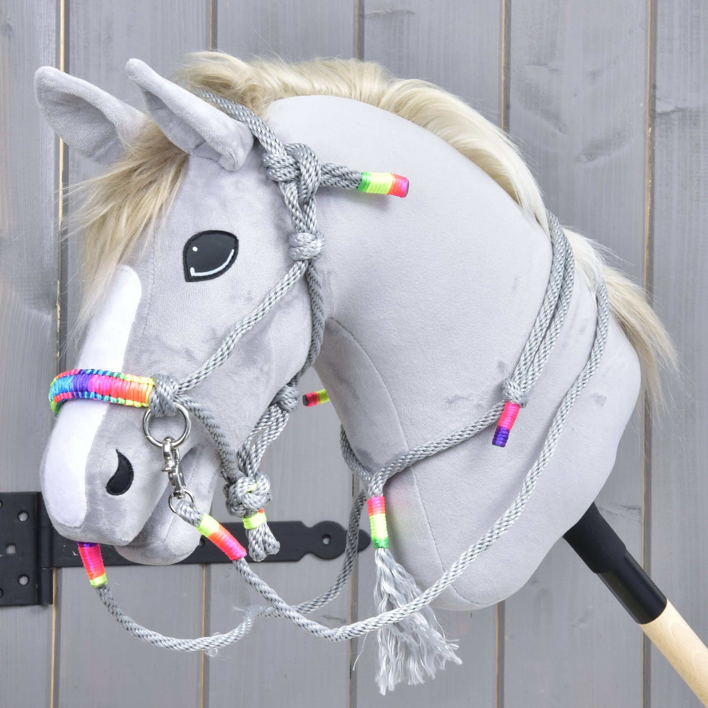 Hobby Horse gris avec liste blanche, nez noir et crinière blonde portant l’ensemble de cordes Manhattan de Hobby Horse LarDen, avec licol en corde grise et rênes à perles colorées, présenté sur fond en bois.