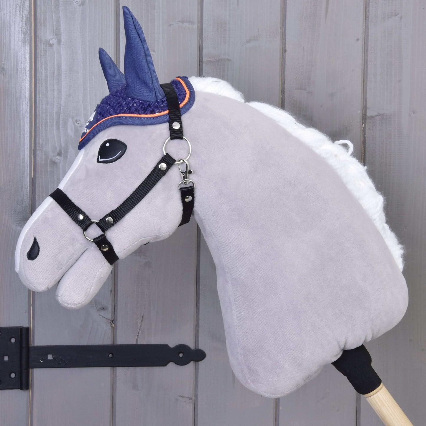 Bonnet d’oreilles au crochet Bleu Sport de Hobby Horse LarDen, avec tête en peluche gris clair, crinière blanche, bride noire et bonnet d’oreilles bleu au crochet sur un bâton en bois, présenté devant un mur en bois clair.