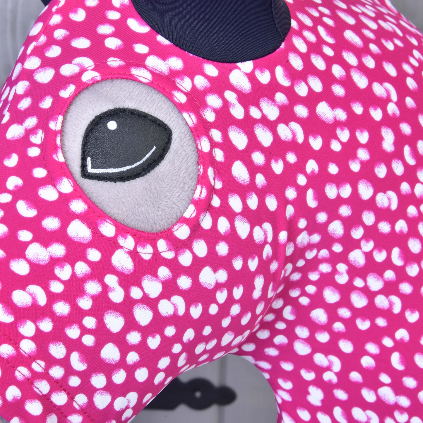 Gros plan sur la combinaison de protection Hobby Horse Minie (Lycra) de Hobby Horse LarDen, une housse lavable rose à taches blanches avec œil gris brodé. Garde votre Hobby Horse propre et élégant. L’arrière-plan flou montre une partie d’un mur gris.