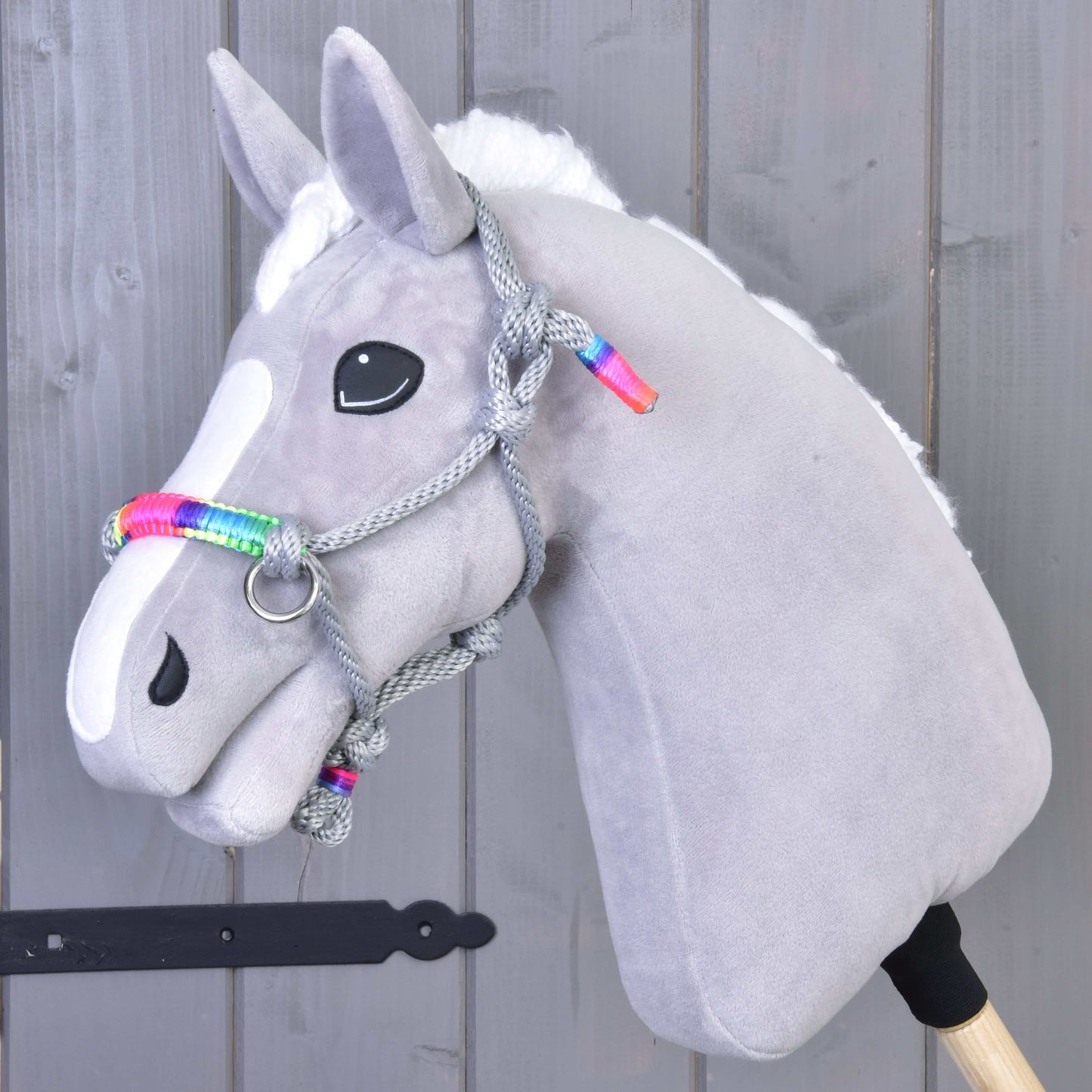 Licou en corde Manhattan de Hobby Horse LarDen, accessoire pour Hobby Horse gris doux avec marque blanche sur le visage, yeux noirs en bouton et licou en corde tressée à accents arc-en-ciel, présenté sur fond en bois gris.