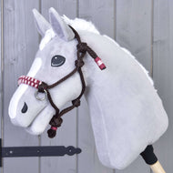 Hobby Horse LarDen Seilhalfter York, ein grauer Plüsch-Hobby Horse Kopf mit weißer Mähne und weißer Nasenzeichnung sowie einem braun geflochtenen Seilhalfter, an einer grauen Holzwand hängend.