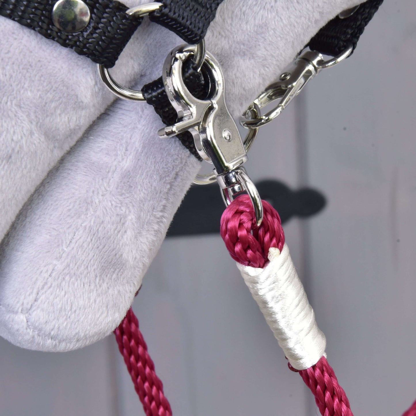 Gros plan sur le mousqueton métallique fixant une corde tressée rose en polypropylène avec ruban blanc à un tissu doux gris clair et à une sangle noire, faisant partie des rênes Czi-czi faites main de Hobby Horse LarDen.