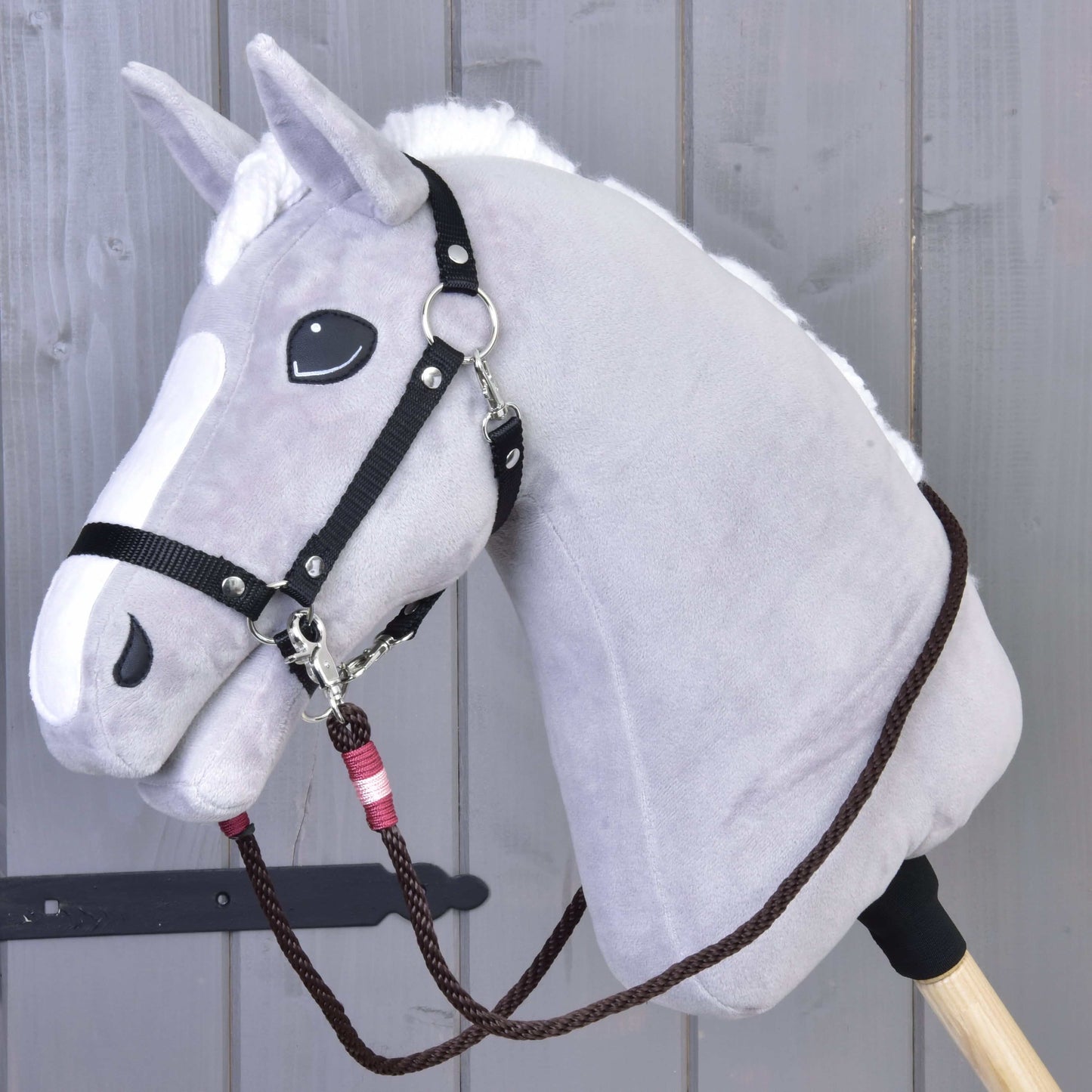 Zügel York von Hobby Horse LarDen an einem grauen Plüsch-Hobby Horse mit weißer Mähne und weißer Schnauze, schwarzen handgefertigten Zügeln aus Polypropylenseil und Holzstab vor Holzpaneelen