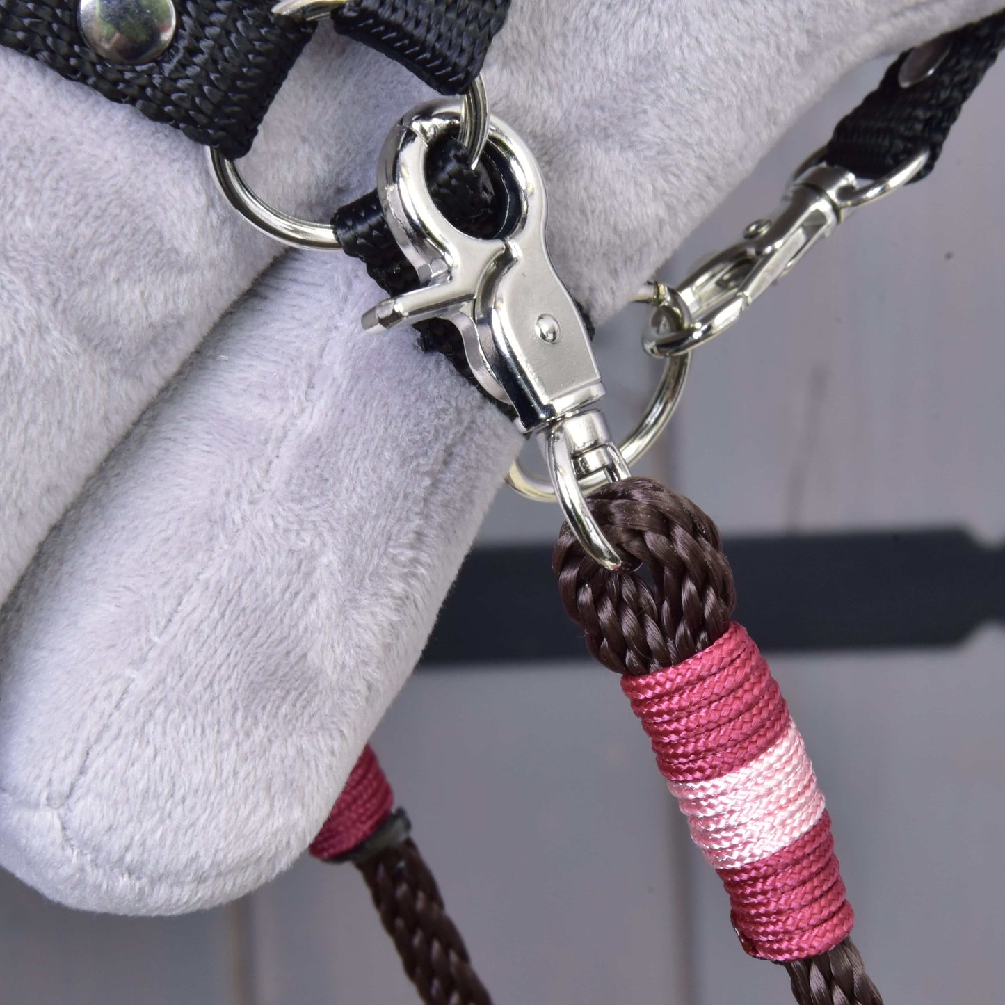 Nahaufnahme eines Metallclips, der Zügel York von Hobby Horse LarDen, braun und pink geflochtenes Polypropylenseil, an einem schwarzen Stoffriemen auf einer weichen hellgrauen Oberfläche befestigt, vermutlich Teil von Hobby Horse Zaumzeug