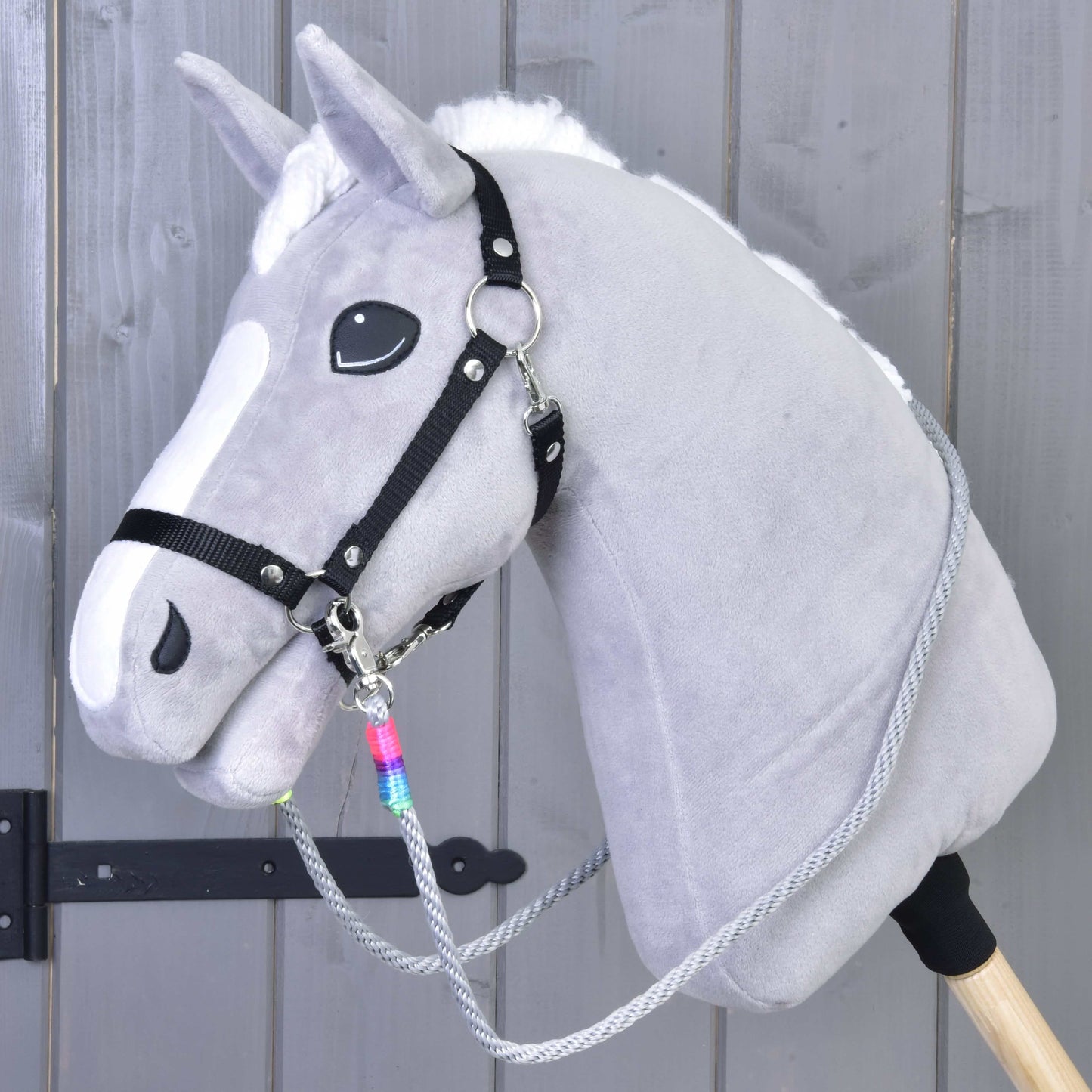 Rênes Manhattan de Hobby Horse LarDen sur un Hobby Horse gris avec crinière blanche, licol noir, rênes faites main et bâton en bois, présenté devant un mur en bois gris.