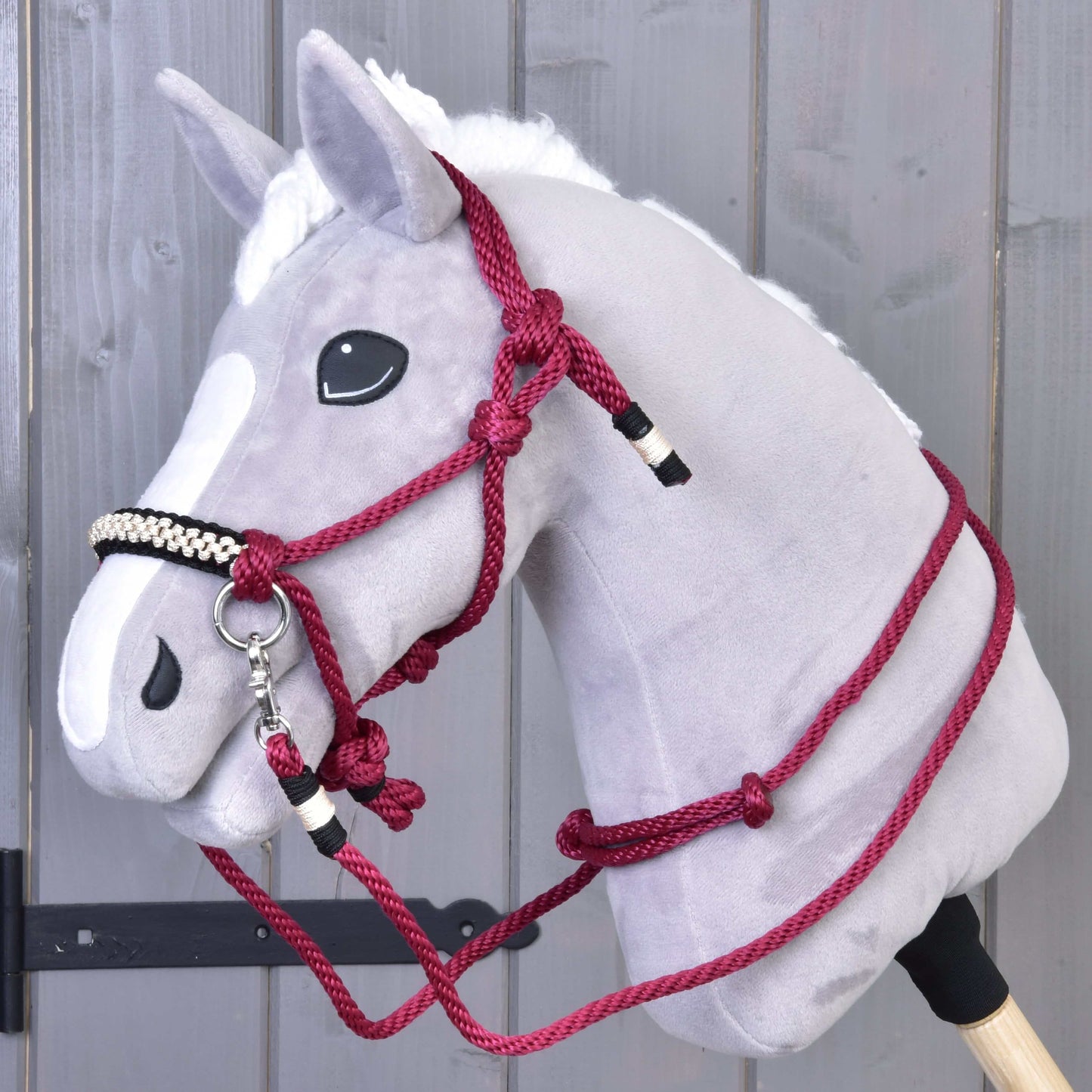 Hobby Horse LarDen Seilset Burgundy zeigt ein graues Plüsch-Hobby Horse mit weißer Mähne, schwarzer Nase und bordeauxrotem Seilhalfter, auf einem Holzstab montiert und vor einer grauen Holzwand.