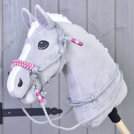 Ensemble de corde Rosie de Hobby Horse LarDen, hobby horse gris en peluche avec liste blanche, yeux noirs et bride rose et grise avec rênes sur un bâton en bois, présenté devant un mur en bois gris clair.