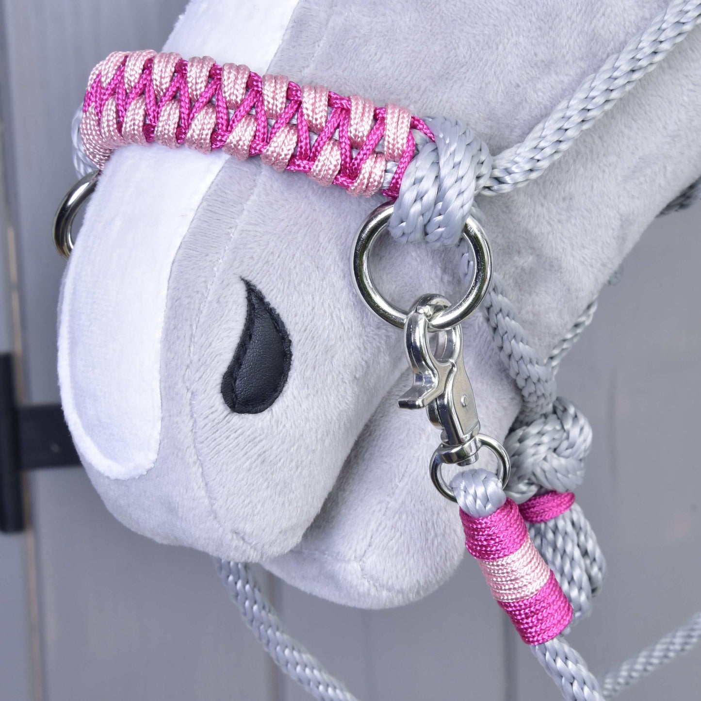 Gros plan sur la tête grise en peluche de Hobby Horse LarDen avec l’Ensemble de corde Rosie : licol et longe tressés roses et argentés, œil noir brodé et mousqueton en métal, sur fond en bois clair.