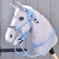 Ein Plüsch-Hobby Horse mit grauem Fell, weißer Mähne und Blesse trägt das Seilset Malibu – ein blaues Seilhalfter mit schwarzen und weißen Akzenten plus passende Zügel – auf einem Holzstab vor einem grauen Holzhintergrund.