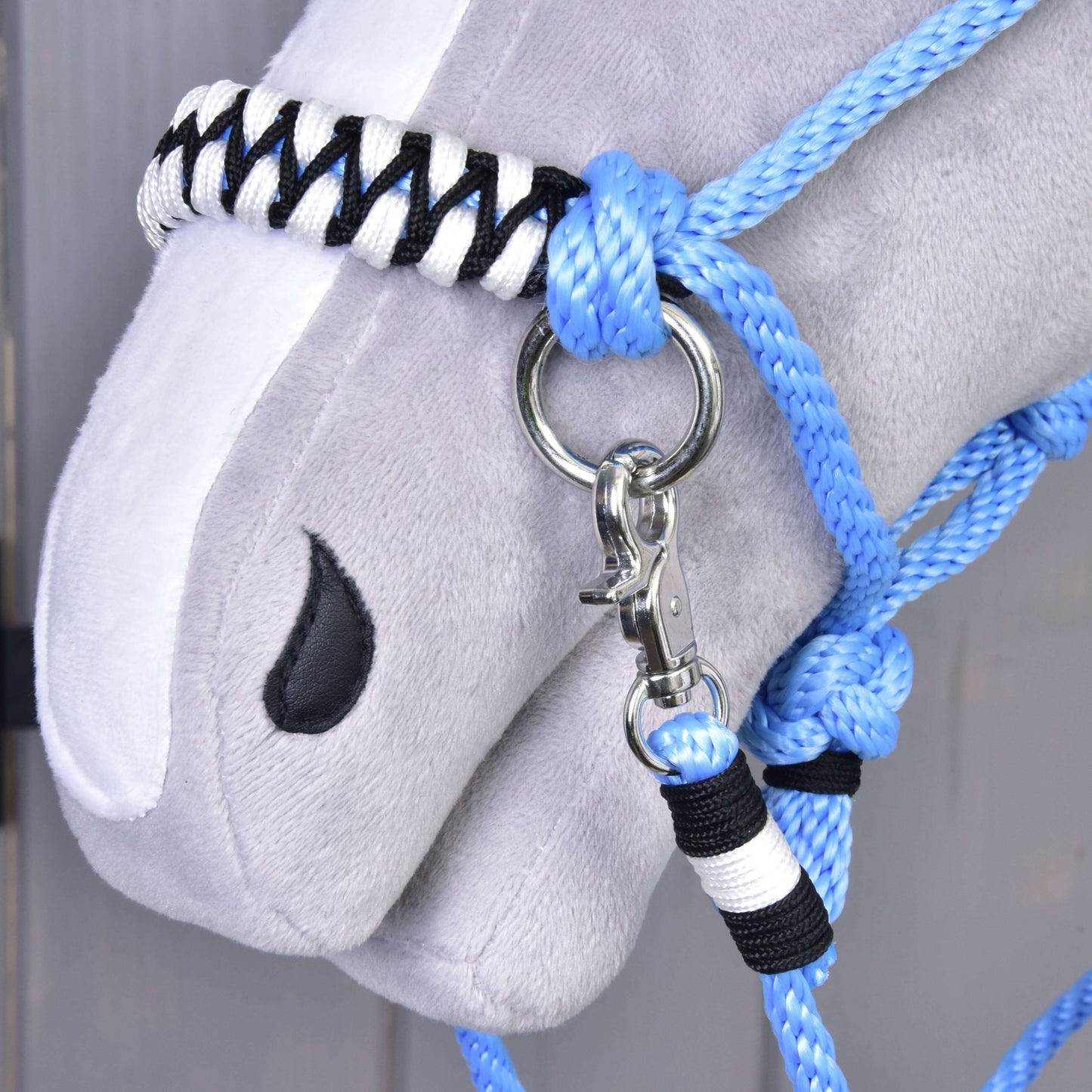 Nahaufnahme des Plüschkopfs eines Hobby Horse mit weißer Blesse, das das blaue Seilhalfter Seilset Malibu mit schwarzen und weißen Akzenten trägt, dazu Zügel, die an einem silbernen Clip und Metallring befestigt sind.