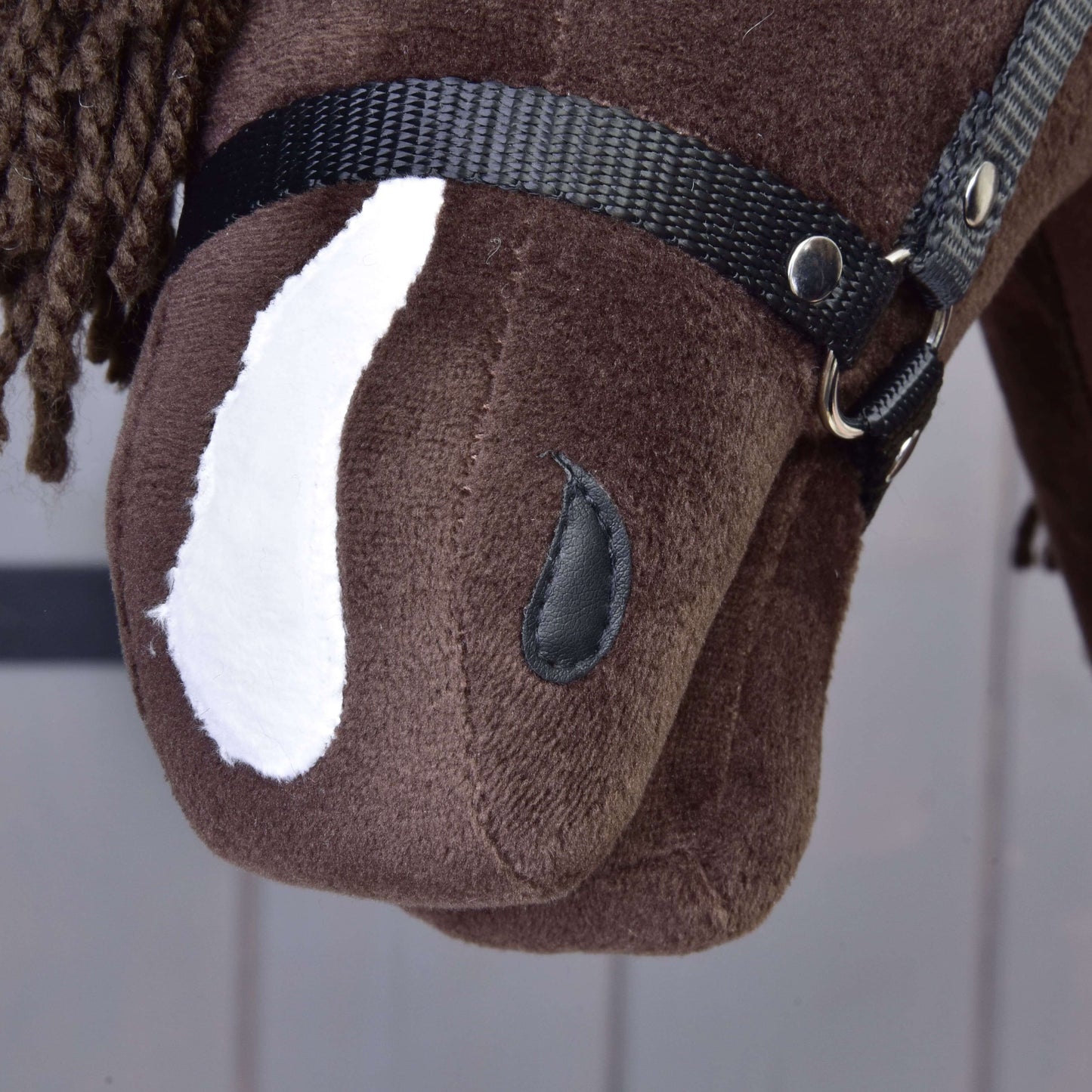 Gros plan sur Hobby Horse Chianti avec licol noir de Hobby Horse LarDen : tête de cheval-jouet en tissu marron avec tache blanche, licol noir, œil en bouton et crinière marron en fil, sur fond clair aux lignes douces.