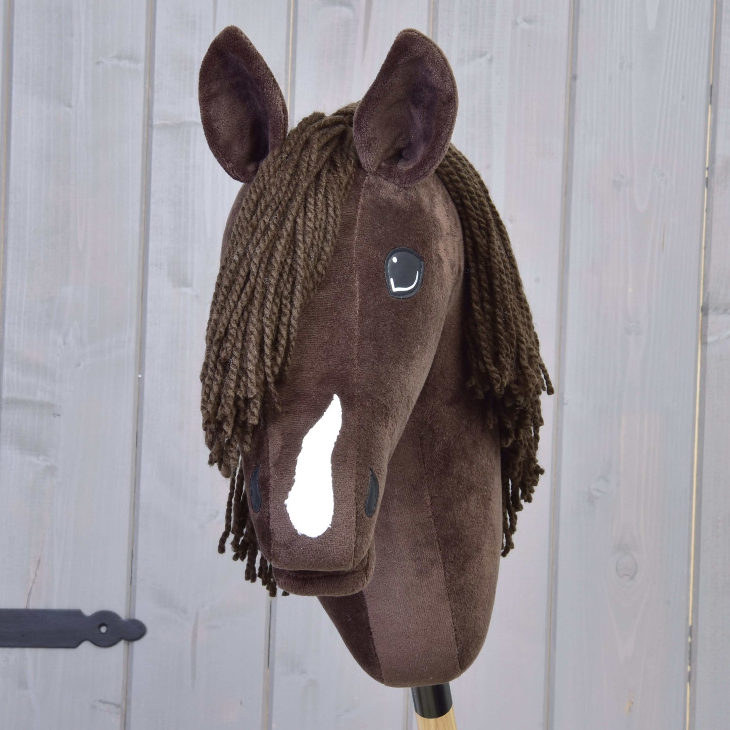 Hobby Horse Chianti avec licol noir de Hobby Horse LarDen, Hobby Horse en peluche marron avec liste blanche, yeux noirs en bouton et crinière marron en fil, inspiré du doux American Quarter Horse, sur fond de panneaux en bois.