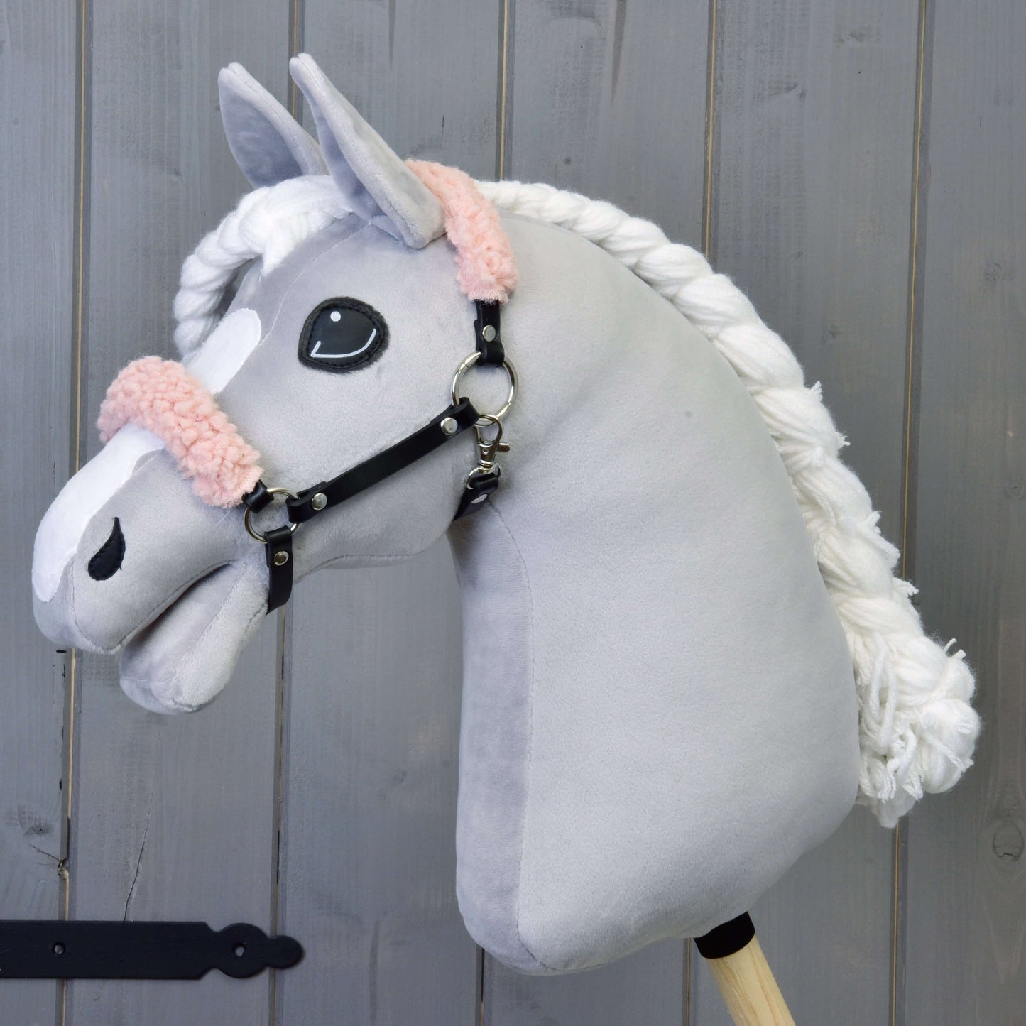 Hobby Horse en peluche Hobby Horse LarDen avec visage gris, crinière blanche et licou noir avec fourrure vieux rose en similicuir, présenté sur fond en bois.