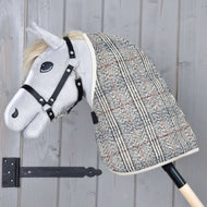A gray plush hobby horse from Hobby Horse LarDen features a black bridle and the Stable Blanket Fine Check, mounted on a wooden stick and set against a light gray wooden wall.