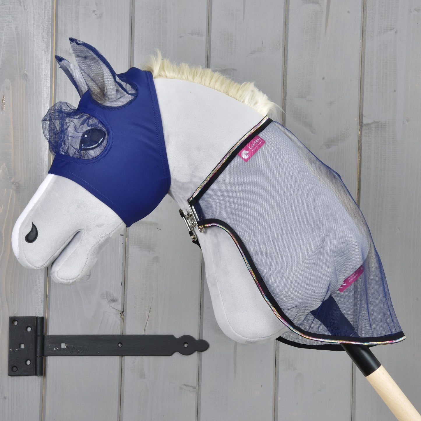 Das Hobby Horse LarDen Set Fly Combo mit einem Plüsch-Hobby Horse mit blauer Fliegenmaske und Fliegendecke aus Netzstoff auf einem Holzstab, vor einer grauen Holzwand, bereit für dein nächstes Abenteuer.
