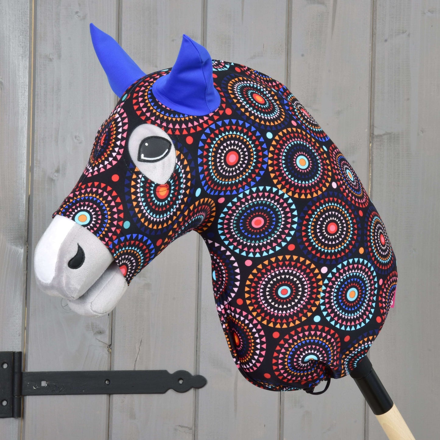 Der Hobby Horse LarDen Hobby Horse Schutzanzug Alice (Lycra) mit einem farbenfroh gemusterten Kopf, blauen Filzohren und ausdrucksstarken Augen auf einem Holzstab, präsentiert vor einer hellgrauen Holzwand.