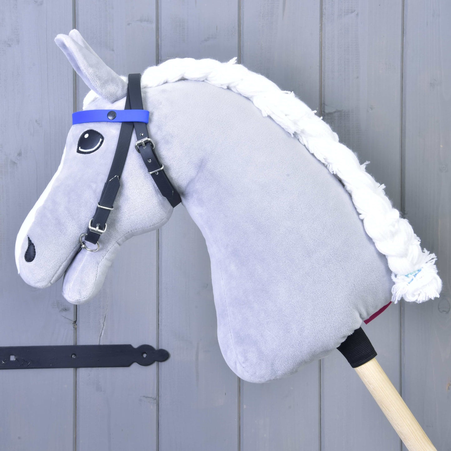 Zaumzeug mit 2 Stirnbändern Blau (Gr. M) von Hobby Horse LarDen an einem plüschigen Hobby Horse mit weißer Garnmähne, befestigt an einem Holzstab und angelehnt an eine graue Holzwand.