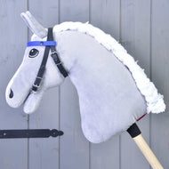 A blue "Bridle with 2 Headbands" (size M) by Hobby Horse LarDen is on a plush hobby horse with a white yarn mane, attached to a wooden stick and resting against a gray wooden wall.