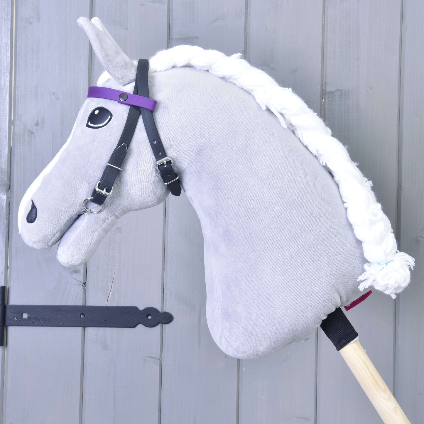 Ein graues Hobby Horse mit weißer Garnmähne und Zaumzeug mit 2 Stirnbändern Lila (Gr. M) von Hobby Horse LarDen, ohne Nasenriemen, auf einem Holzstab vor einer hellgrauen Holzwand.