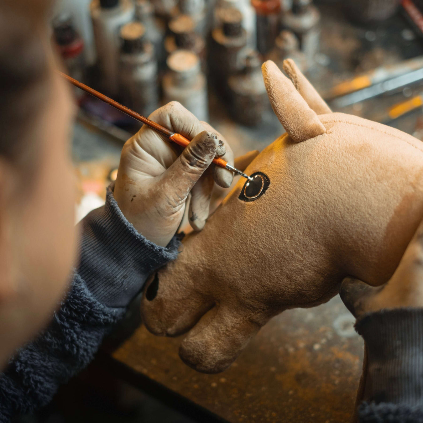 Eine Person mit Handschuhen bemalt mit einem Pinsel sorgfältig das Auge eines braunen plüschigen Hobby Horse-Spielzeugs, während im unscharfen Hintergrund Farbtöpfe zu sehen sind.