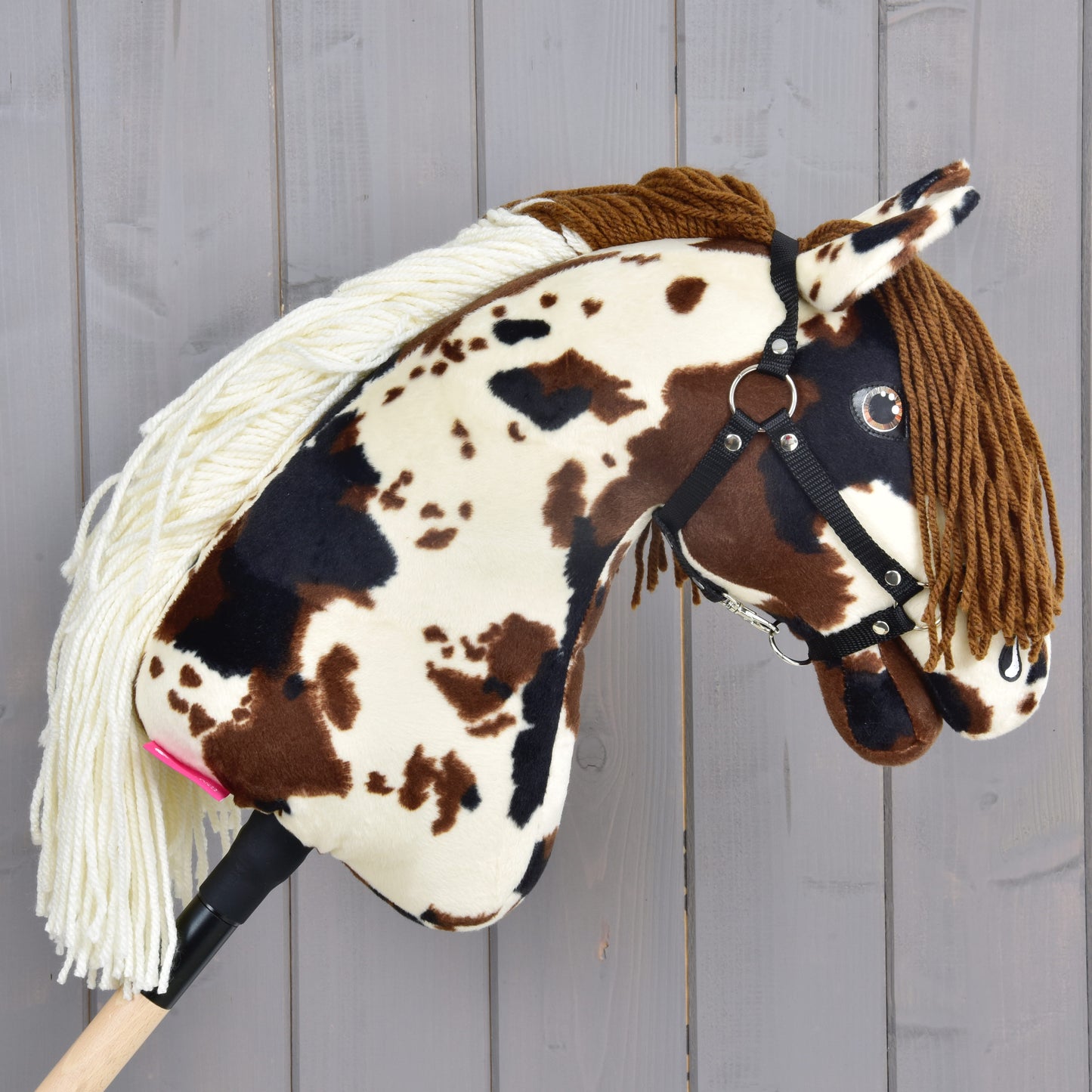 The Hobby Horse LarDen "Hobby Horse Dakota with Black Halter" features a plush brown and white spotted design, white yarn mane, brown forelock, black halter, and wooden stick. Gender-neutral style; shown against a gray wooden background.