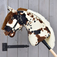 Hobby Horse Dakota mit schwarzem Halfter von Hobby Horse LarDen mit braunen, schwarzen und weißen Flecken, brauner Garnmähne und schwarzem Halfter, auf einem Holzstab vor einer grauen Wand, das Design ist für jedes Geschlecht geeignet.
