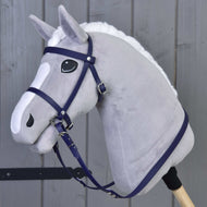 A plush gray hobby horse by Hobby Horse LarDen, featuring a complete bridle with noseband in dark purple, black and white eyes, white mane, and mounted on a wooden stick, stands before a gray wooden wall.