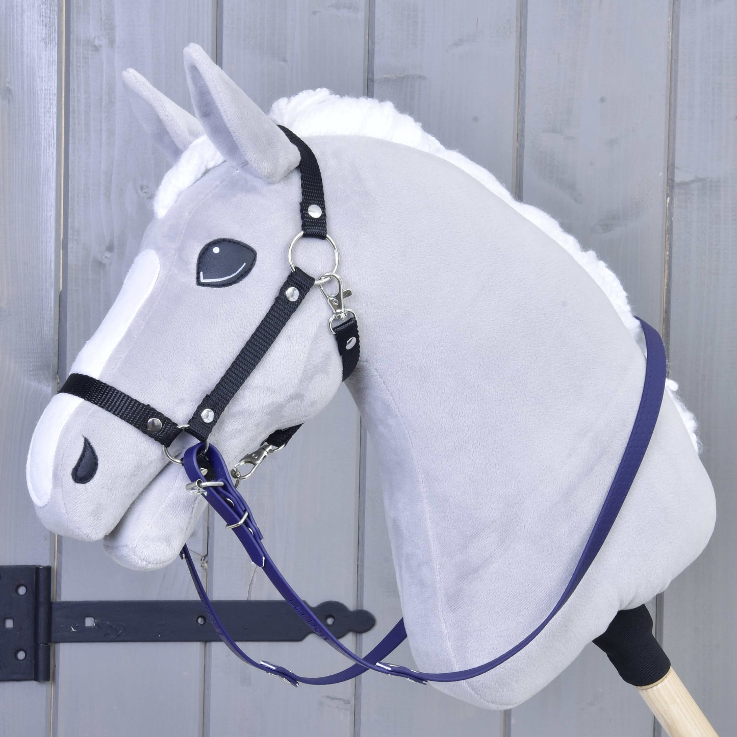 Rênes caoutchoutées violet foncé de Hobby Horse LarDen sur un Hobby Horse gris en peluche avec bride noire, crinière blanche et bâton en bois, en gros plan devant un fond en bois gris clair.