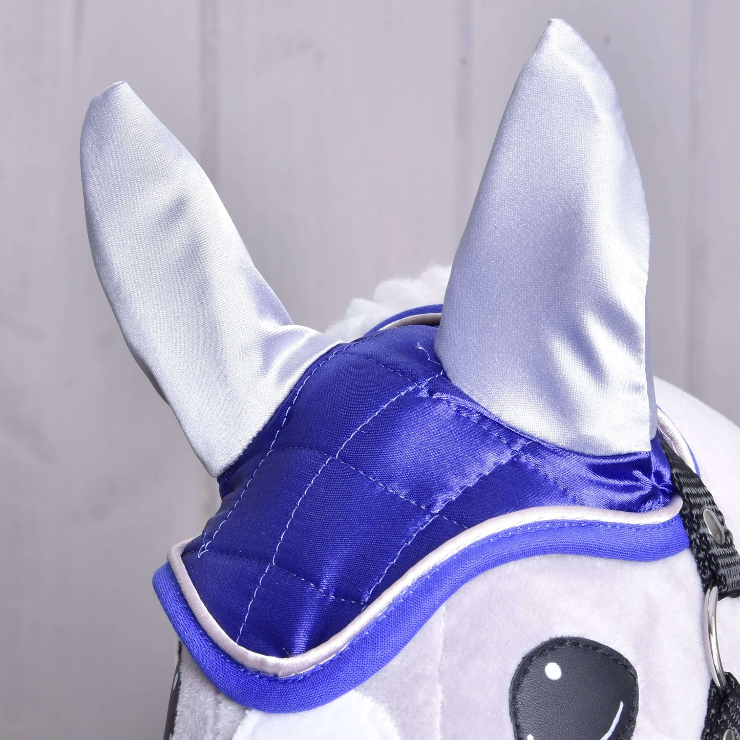 Nahaufnahme der Hobby Horse LarDen Ohrenkappe DeLuxe Blue auf einem Plüsch-Hobby Horse mit glänzendem blauem und silbernem Stoff zum Insektenschutz, großen spitzen Ohren und schwarz-weiß gestickten Augen vor hellem Hintergrund.