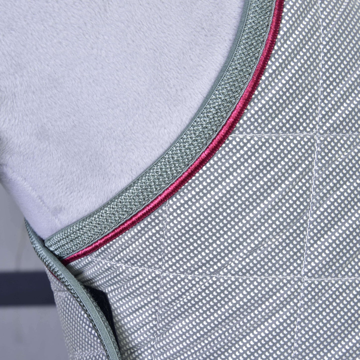 Gros plan sur la Couverture Argent Élégance de Hobby Horse LarDen : couverture d’entraînement grise à motif losanges avec bordure vert foncé et accents rouges. Le fond doux gris clair met en valeur ses coutures détaillées pour une protection optimale du cheval.