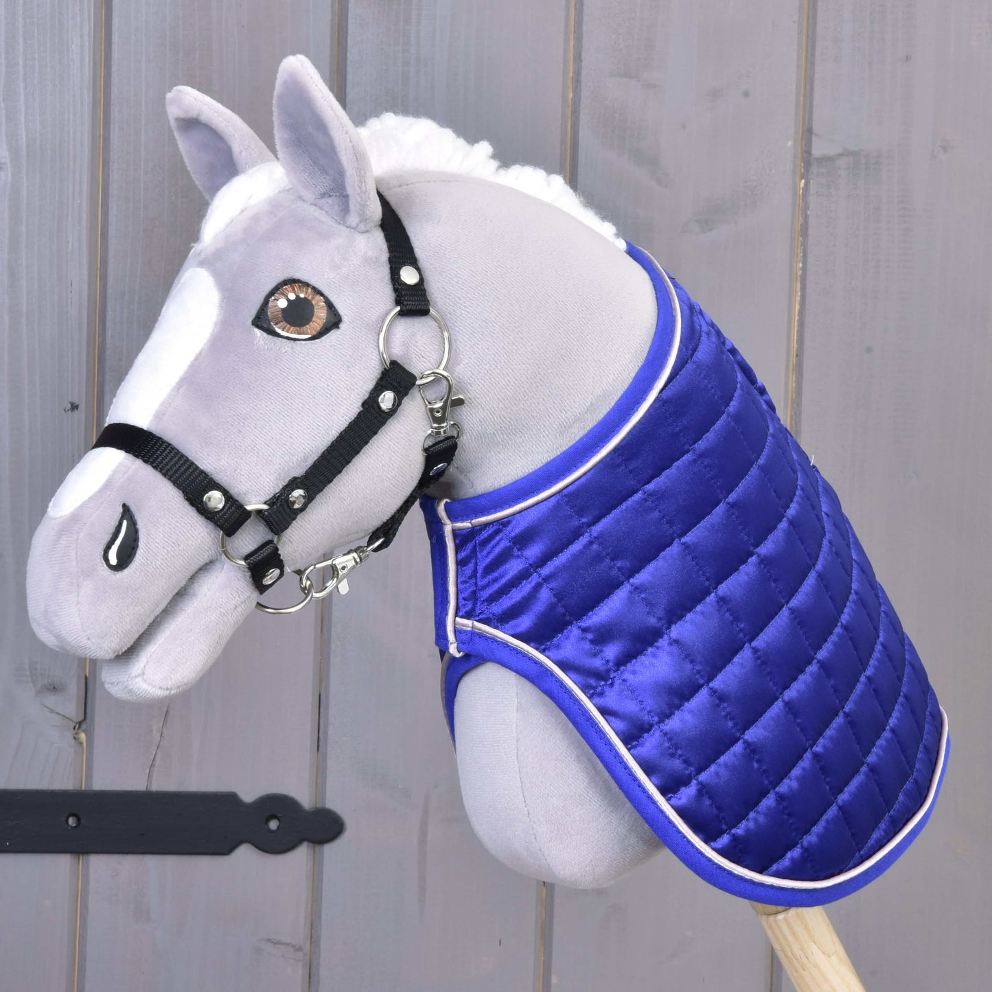 Ein Plüsch-Hobby Horse mit weißer Mähne, schwarzem Halfter für Hobby Horse und der Hobby Horse LarDen Decke DeLuxe Blue doppelseitig für zusätzlichen Schutz ist an einem Holzstab vor grauem Holzbackground befestigt.