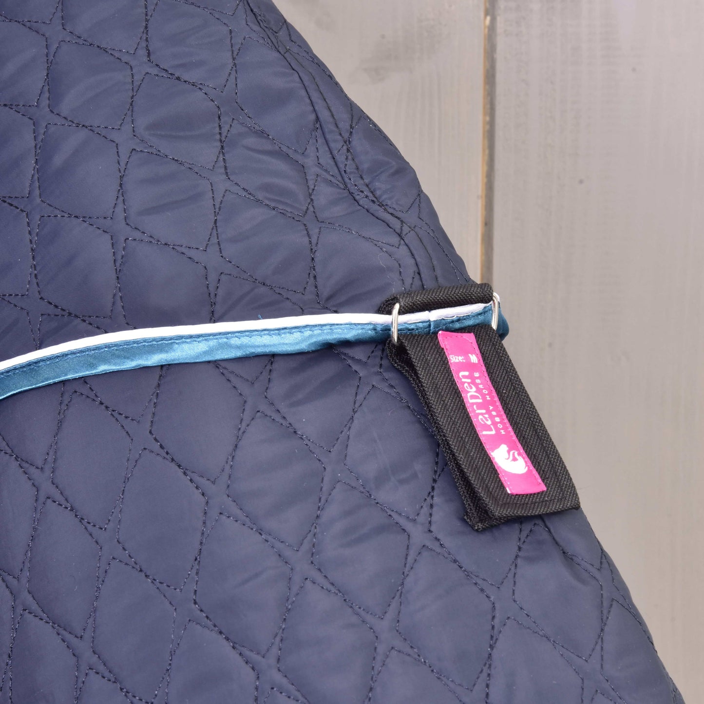 Nahaufnahme der Hobby Horse LarDen Decke mit abnehmbarem Halsteil Mandarin in Dunkelblau mit hellblauem Riemen mit Metallring und einem schwarz-pinken Etikett mit Text und Pferdegrafik, auf hellem Holzuntergrund.
