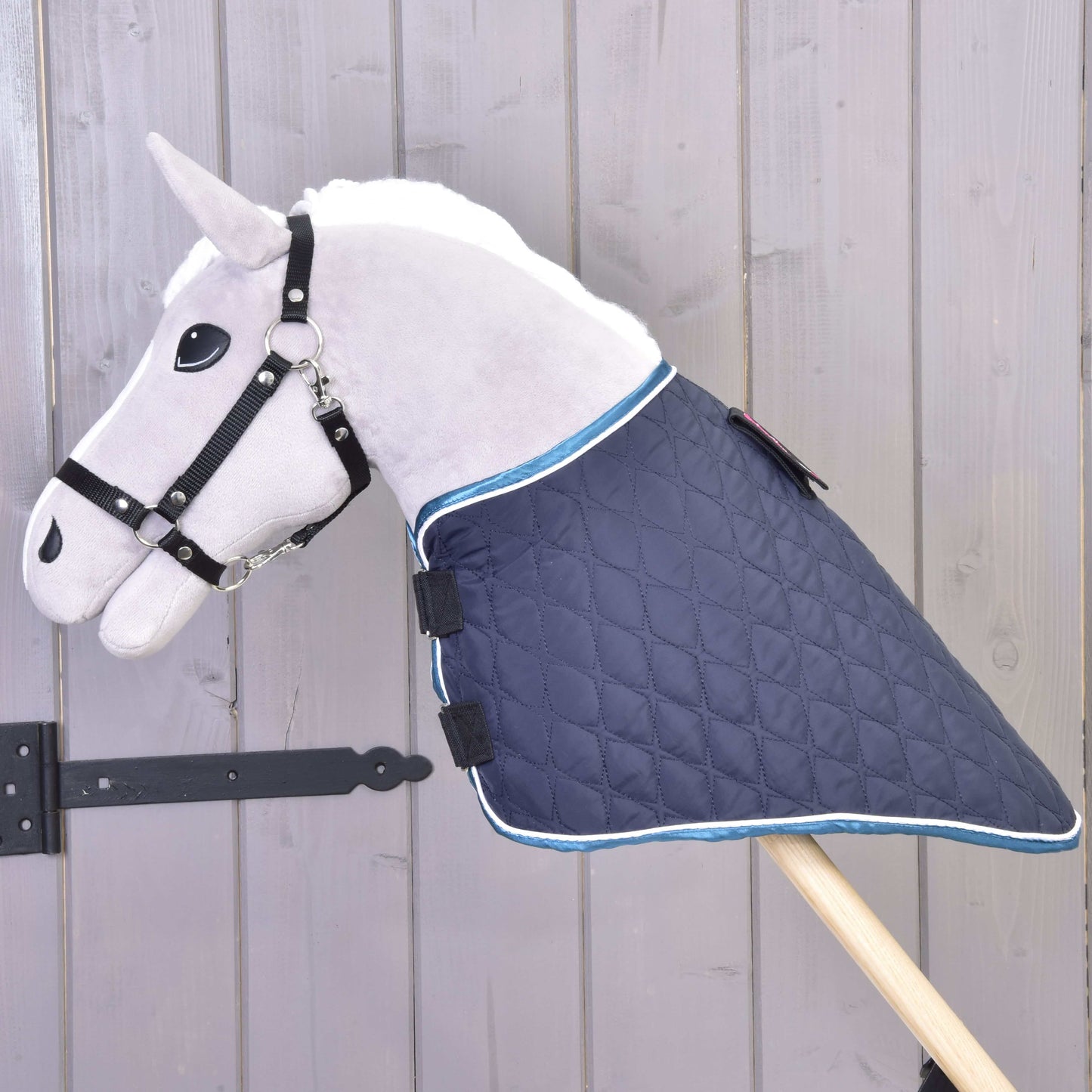 Ein Plüsch-Hobby Horse LarDen mit weißer Mähne, das die Decke mit abnehmbarem Halsteil Mandarin trägt, lehnt an einer hellen Holzwand.