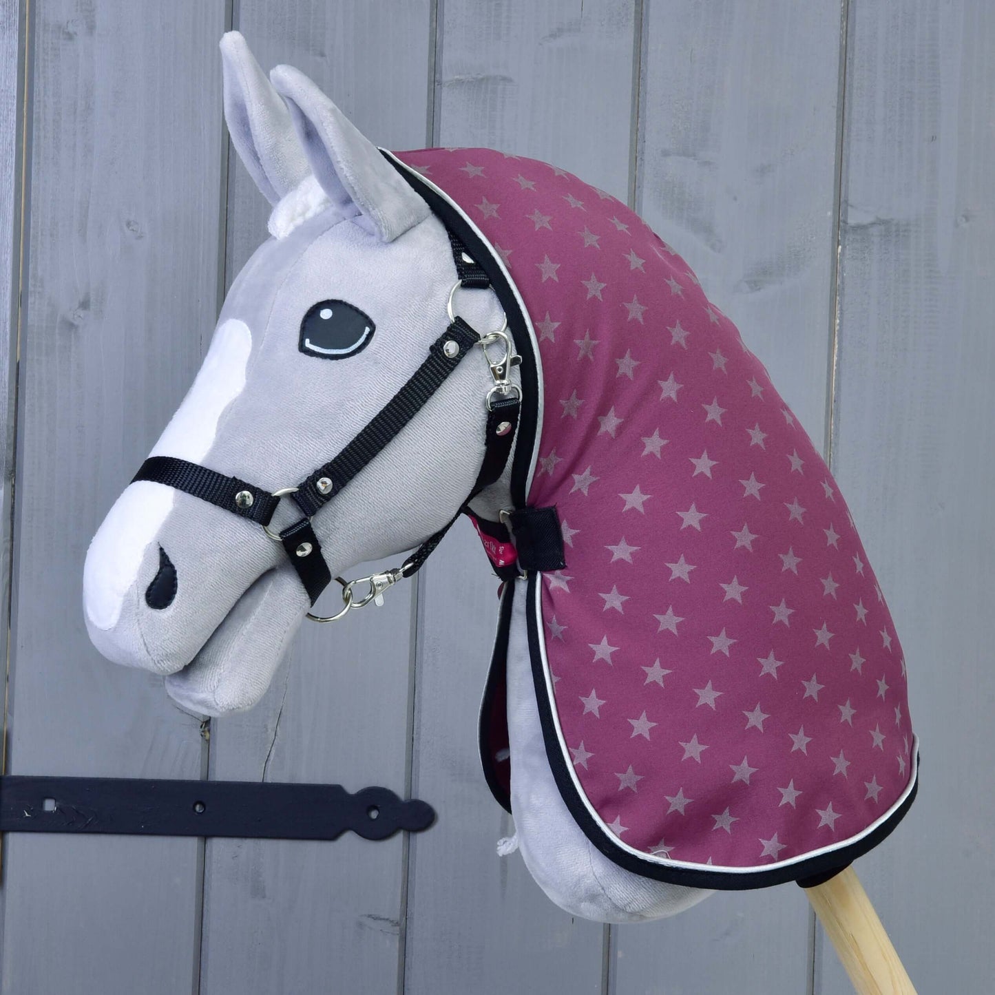 Hobby Horse gris de Hobby Horse LarDen portant une bride noire, une cagoule bordeaux à motif étoilé et la couverture d'écurie violette, devant un fond en bois gris. Il présente une liste blanche et des yeux noirs en bouton.
